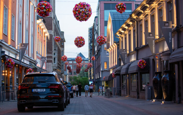 Gata med upplysta fasader, parkerade bilar, gående människor och pynt i form av blom-bollar som hänger i luften